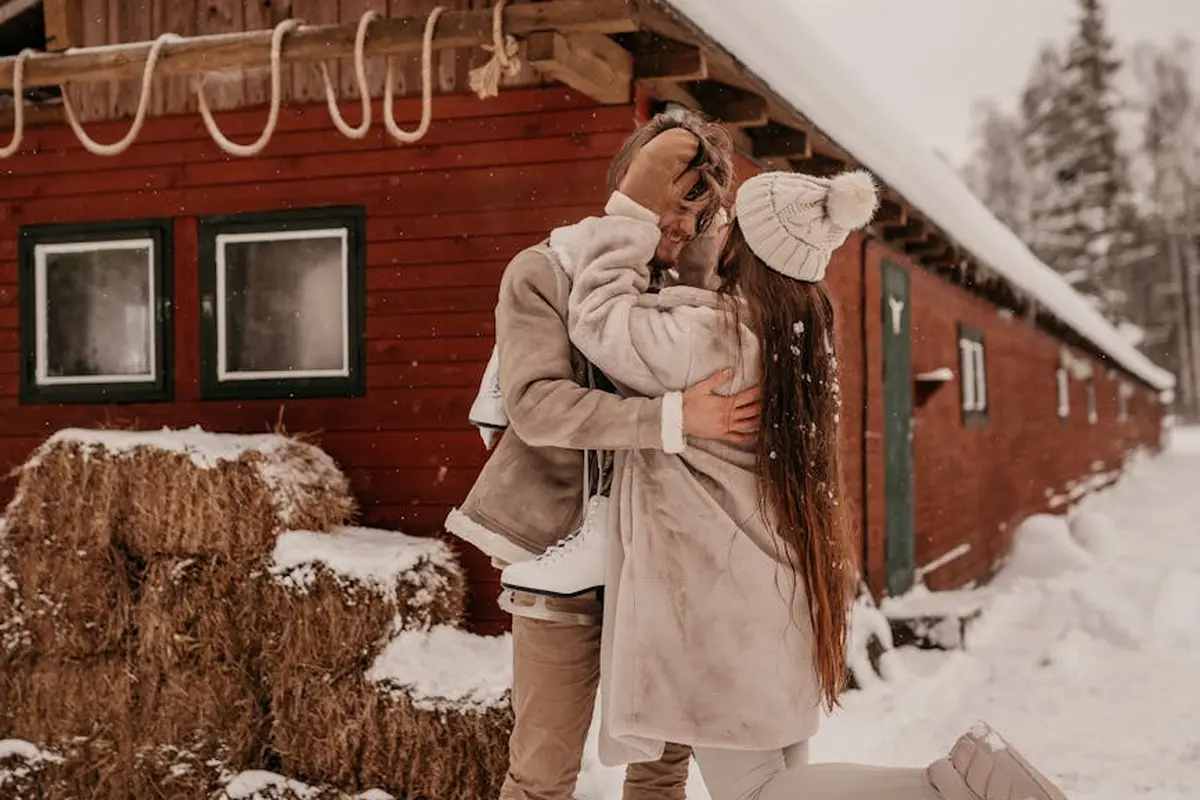 Two people in winter coats embrace and kiss in a snowy rural setting beside a red wooden barn.