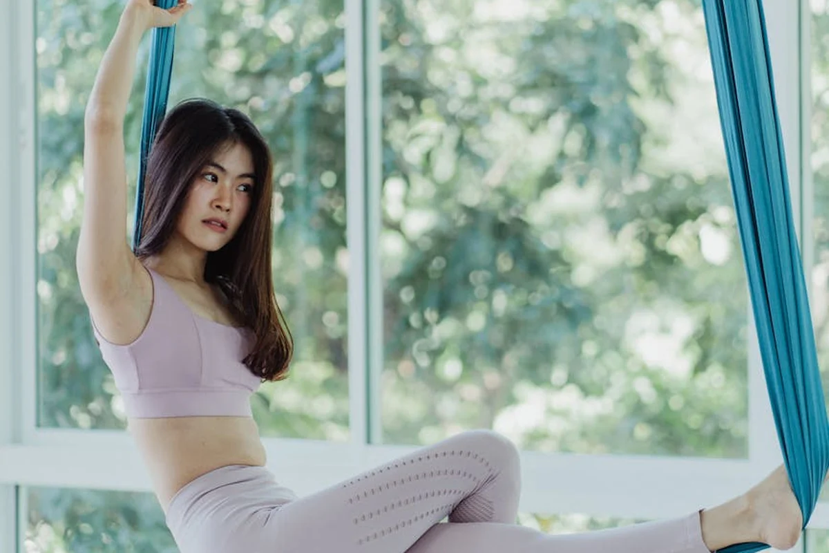 Woman in lavender athletic wear sits on blue silk fabric suspended in a sunlit studio.