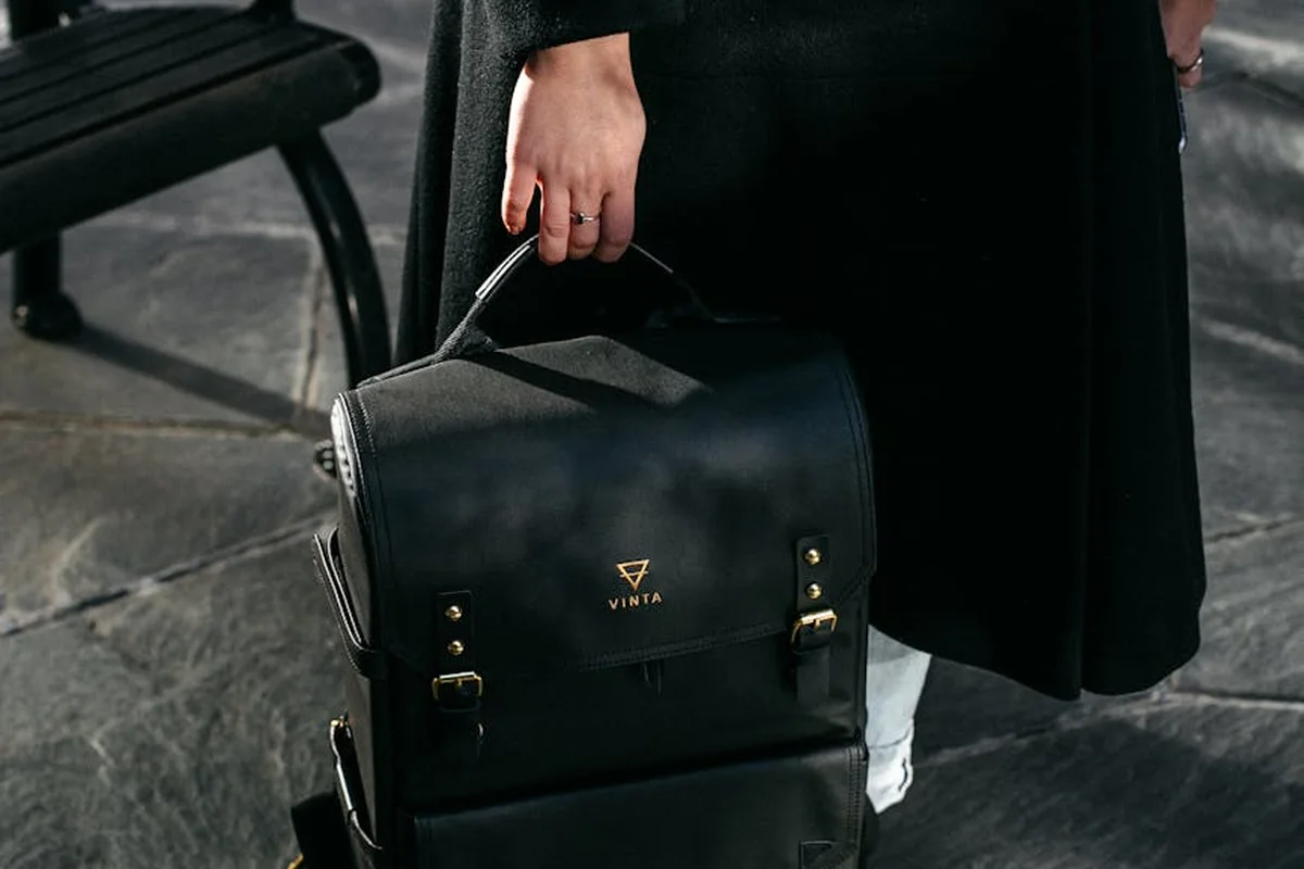 A black leather-looking backpack with metal buckles is held by a person wearing a dark coat, standing on stone pavement.