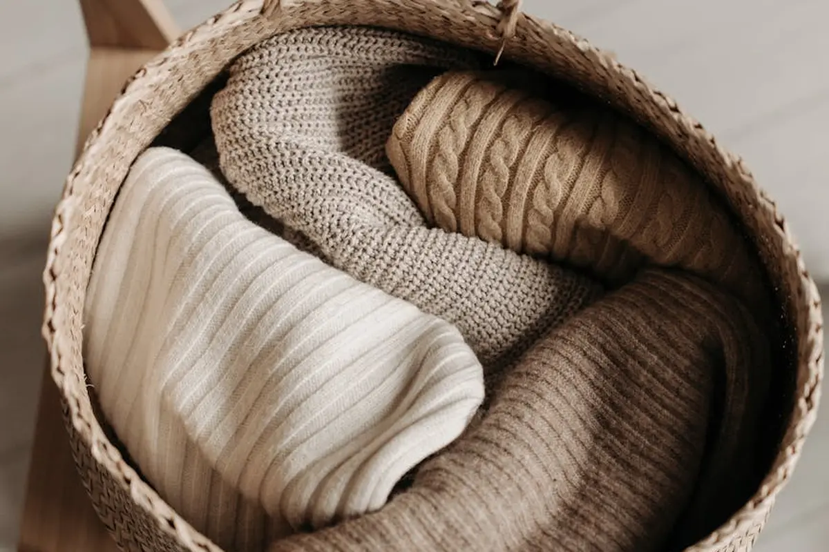 Wicker basket filled with neatly folded cream, beige, and brown fabrics, suggesting textiles suitable for adhesive projects.