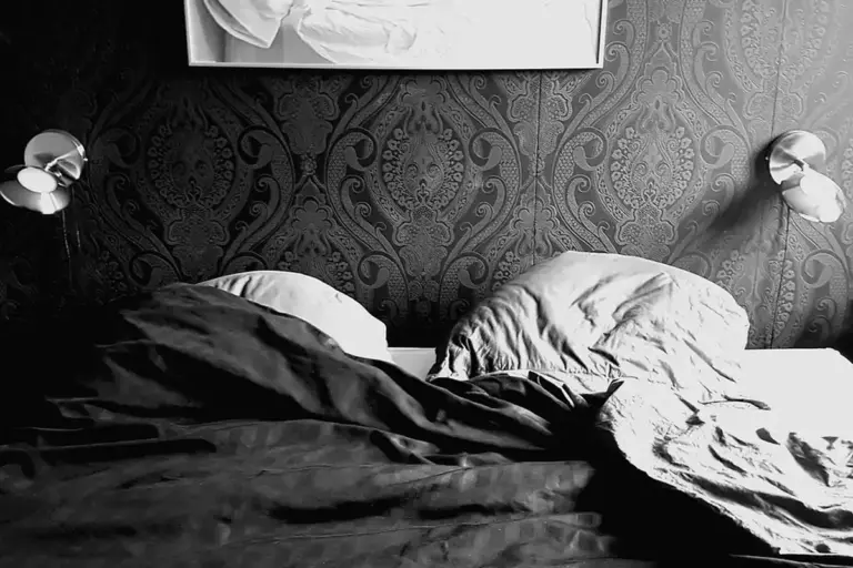 Black-and-white photo of a rumpled bed with crumpled sheets, flanked by two wall lamps and ornate wallpaper.