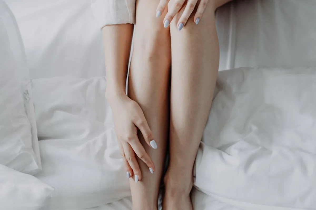 Close-up of a person's legs and hands resting on white bed sheets.