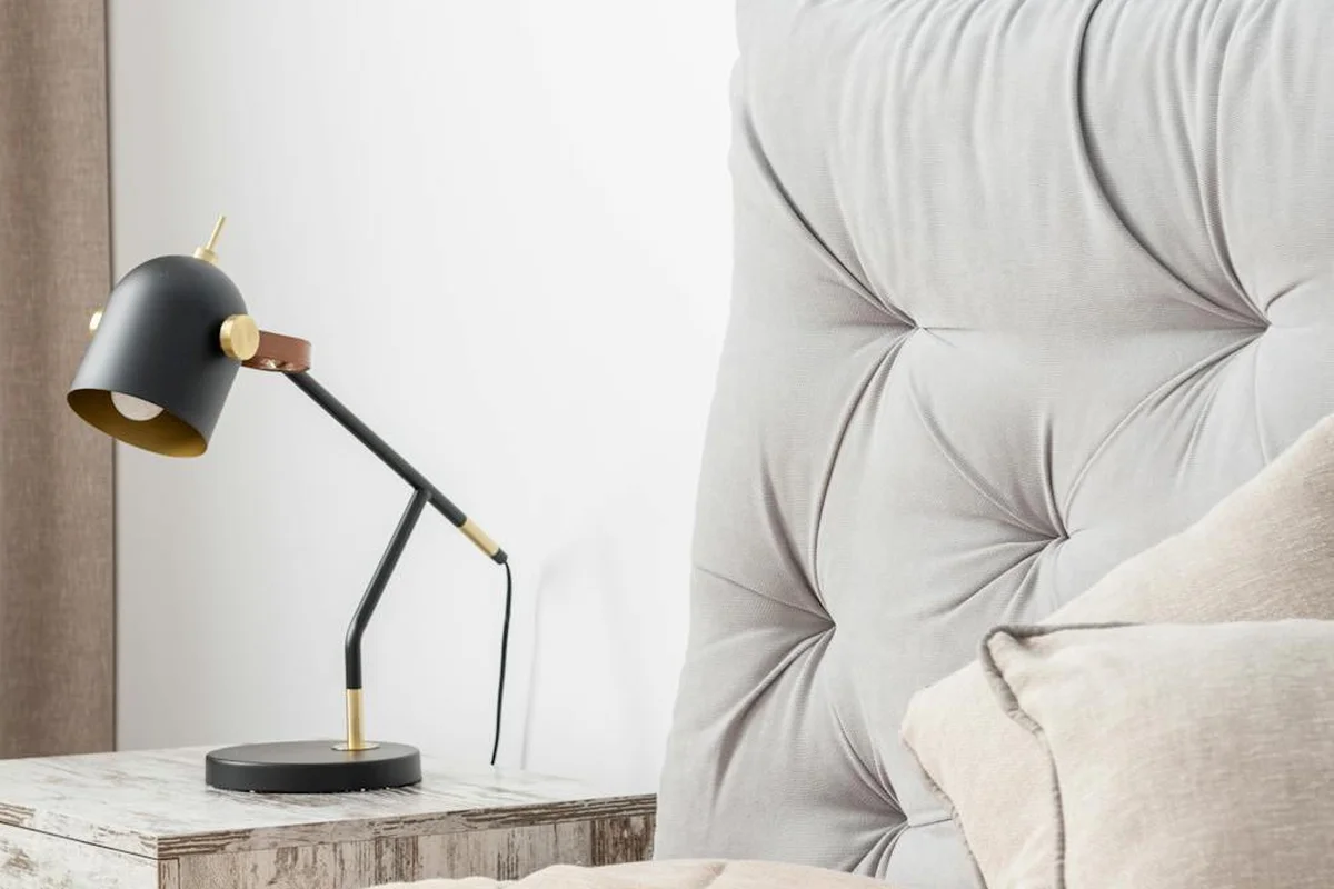A modern bedroom scene featuring a black and brass desk lamp on a weathered wooden bedside table beside a tufted beige headboard and soft bedding.