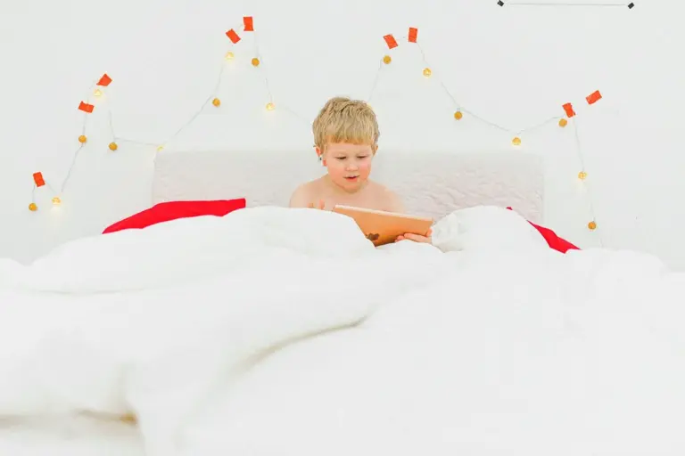 A young child sits up in a white bed, using a tablet, with string lights on the wall behind.