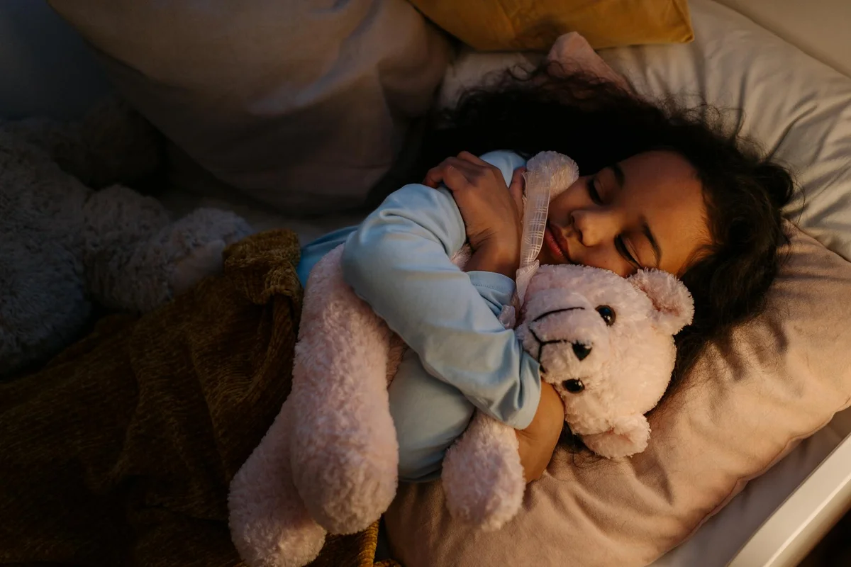 Child sleeping in bed, hugging a light-colored teddy bear.