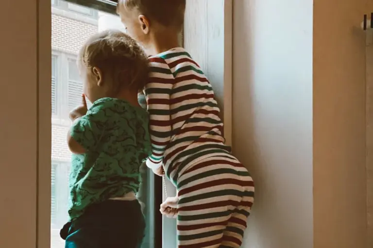 Two children in striped and green pajamas standing by a window, looking outside.