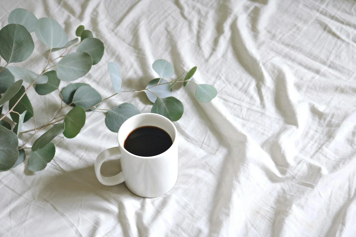 White ceramic cup of black coffee resting on a light linen surface with sprigs of eucalyptus.