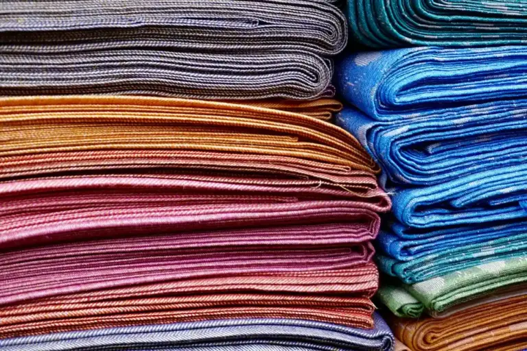 Stacks of folded fabrics in various colors including cotton, wool, and polyester.