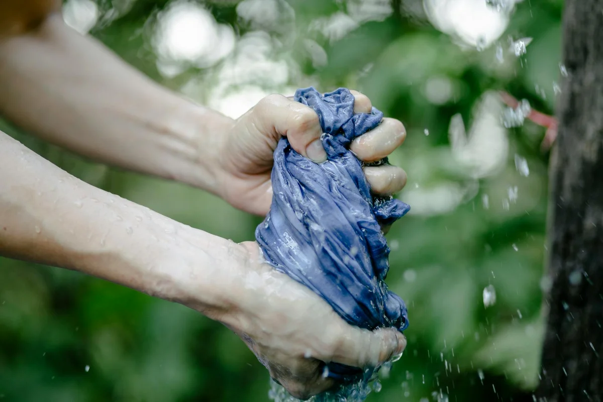 Hands wringing a wet blue fabric (polyester-spandex blend) under running water, with droplets and a green background.