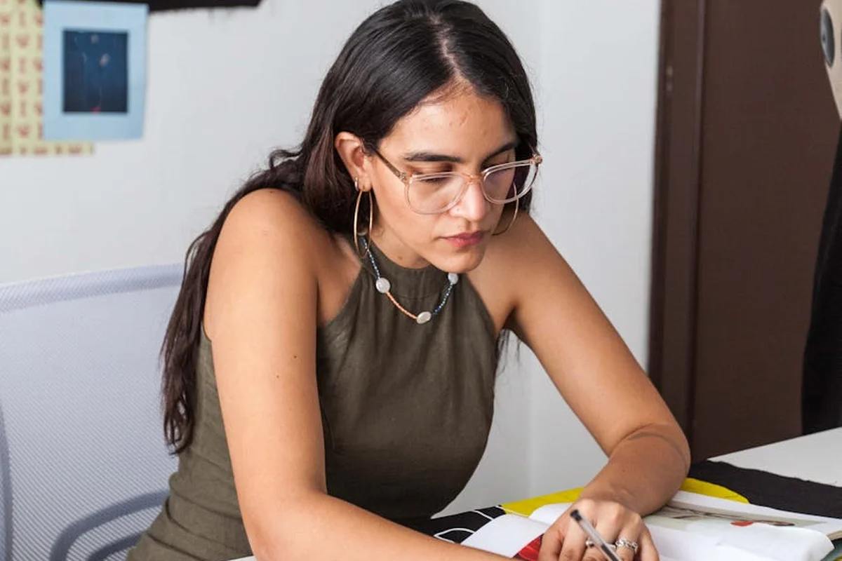 Woman wearing glasses sits at a desk, concentrating on a fabric project.