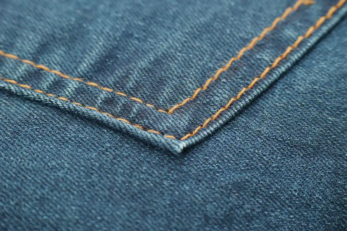 Close-up view of blue denim fabric with orange topstitching along a seam.