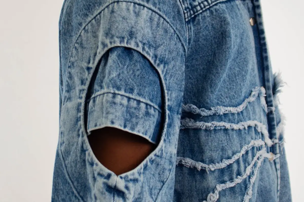 Close-up of a light-washed denim jacket with distressed rips and a sleeve cutout, highlighting textured fabric.