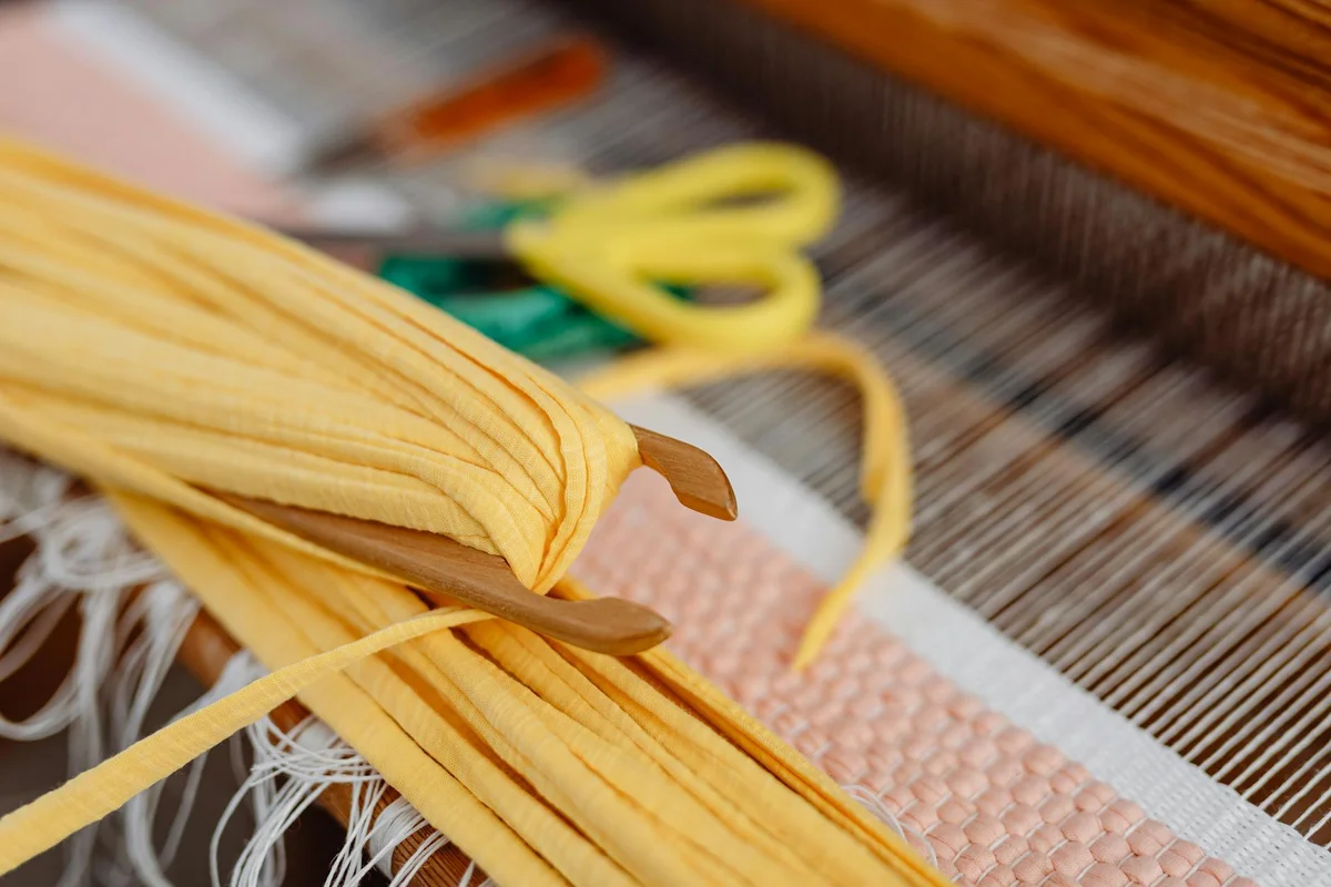 Close-up of yellow flat ribbons on a loom with woven fabric and fringe threads visible, illustrating weaving structure.