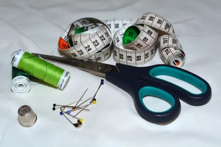 Scissors, a measuring tape, pins, a spool of green thread, a needle and thimble laid on white fabric, showcasing traditional sewing tools.