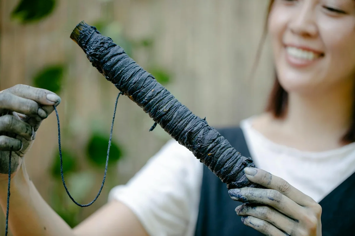 Smiling person wearing gloves holds a tightly wrapped fabric dye bundle outdoors.