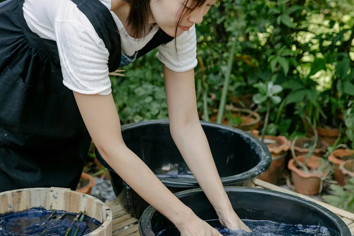 Person wearing a white shirt and dark apron leaning over a large dye tub outdoors, hands immersed in blue dye, with dye containers and potted plants in the background