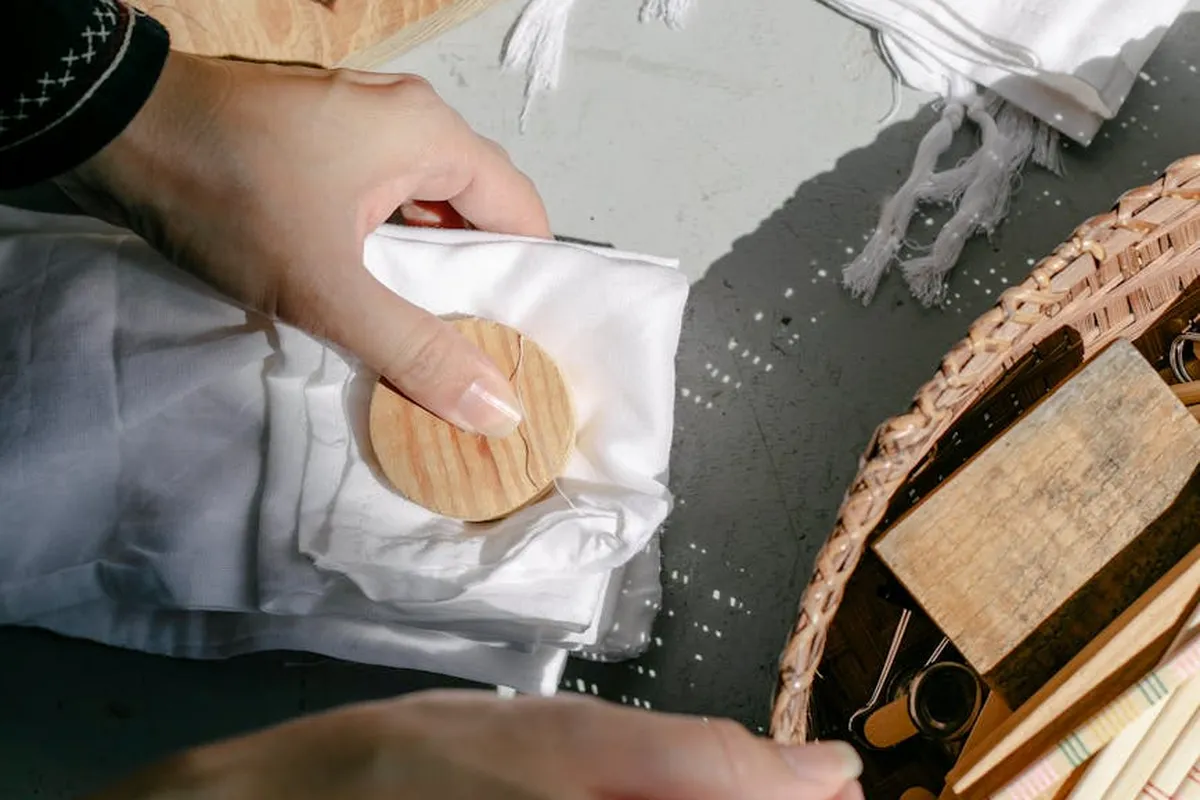 Close-up of hands pressing white fabric with a wooden disc, part of the scouring prep for natural dyeing; a wicker basket with dye tools sits nearby.