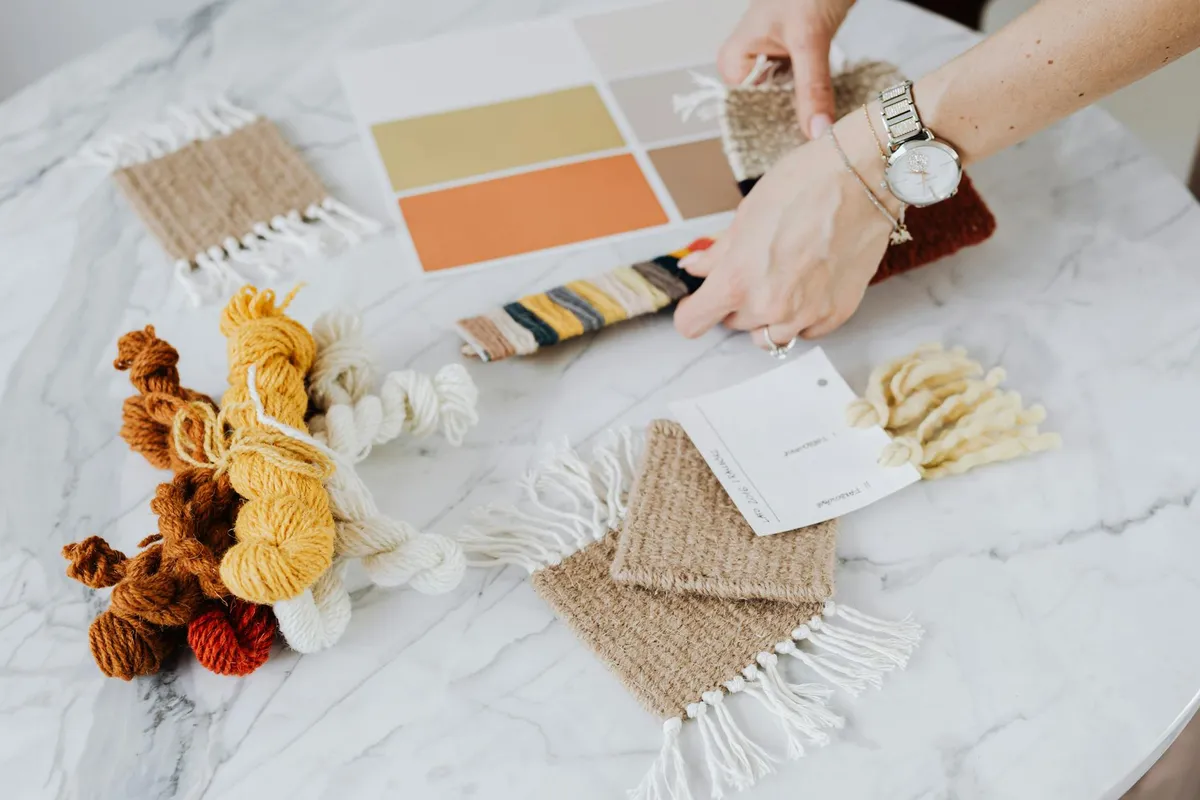 Hands arranging fabric swatches and a color palette on a marble table, with yarn skeins nearby