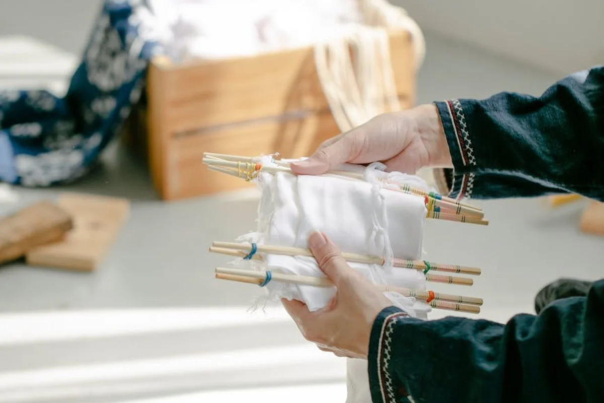 Close-up of hands weaving white fabric on a loom with wooden dowels.