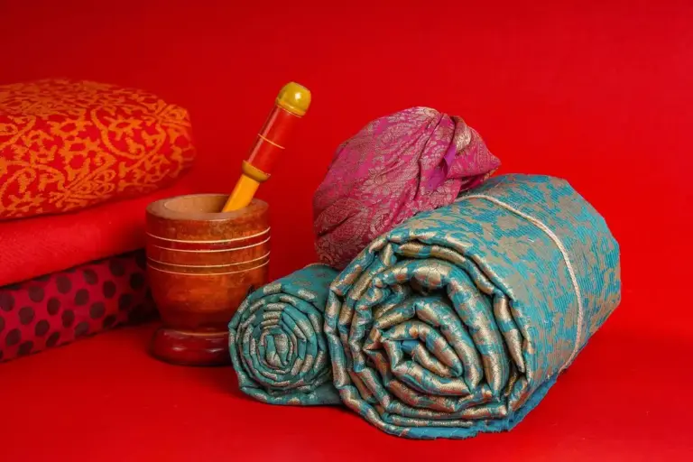 Assorted fabric bolts rolled and stacked on a bright red background, including turquoise patterned fabrics and a wooden thread spool nearby.