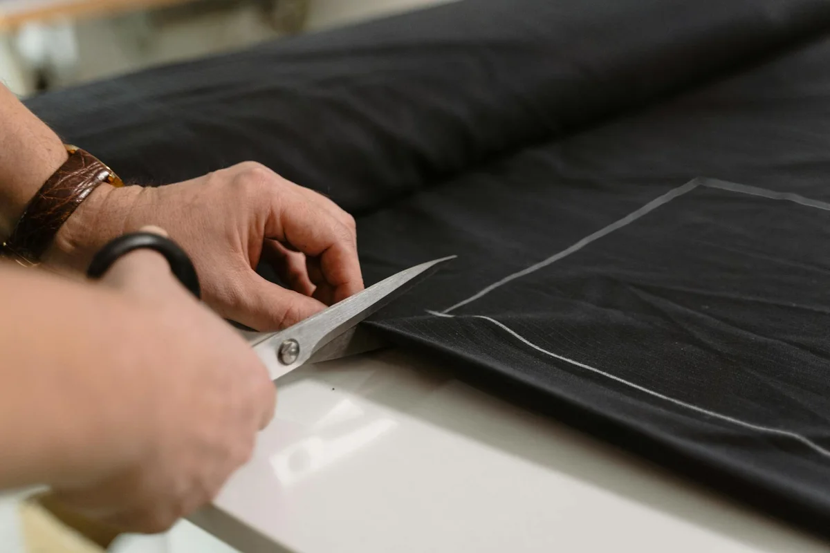 Close-up of hands cutting a black fabric with scissors, showing the fabric's texture and edge lines.