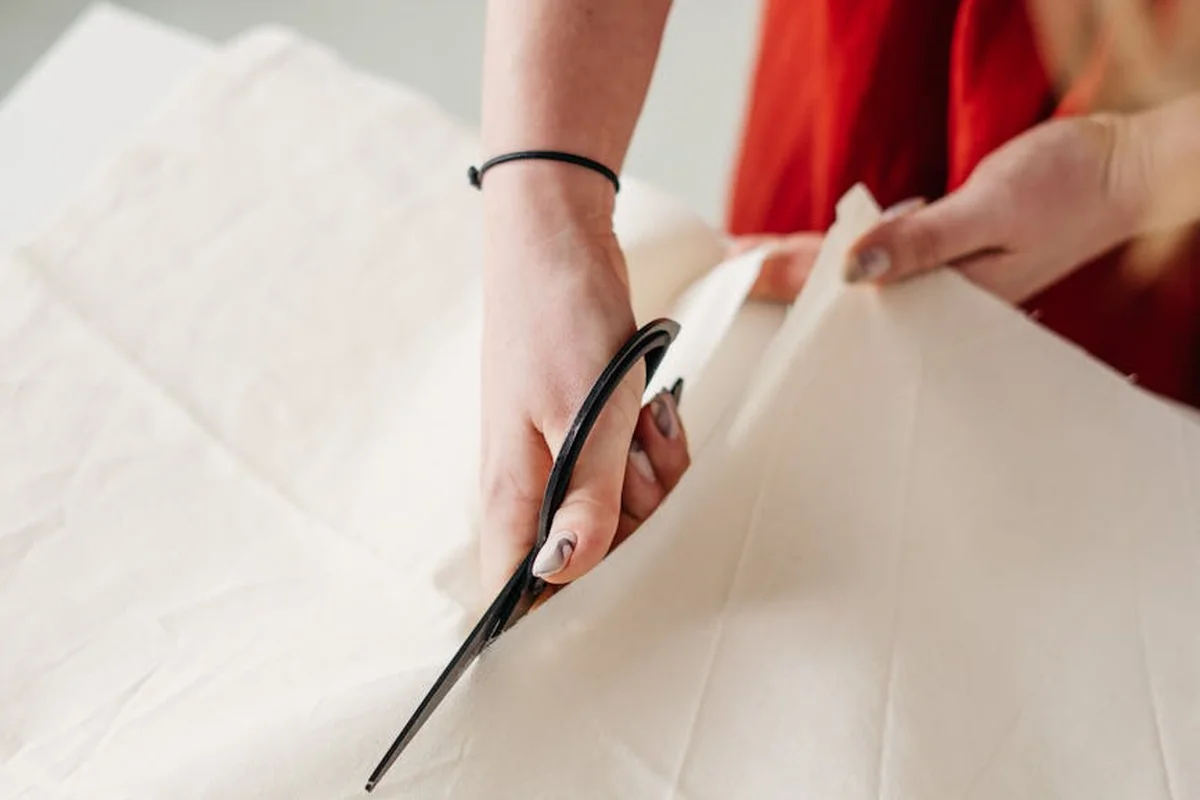 Close-up of hands cutting white fabric with scissors, with a hint of red clothing in the background
