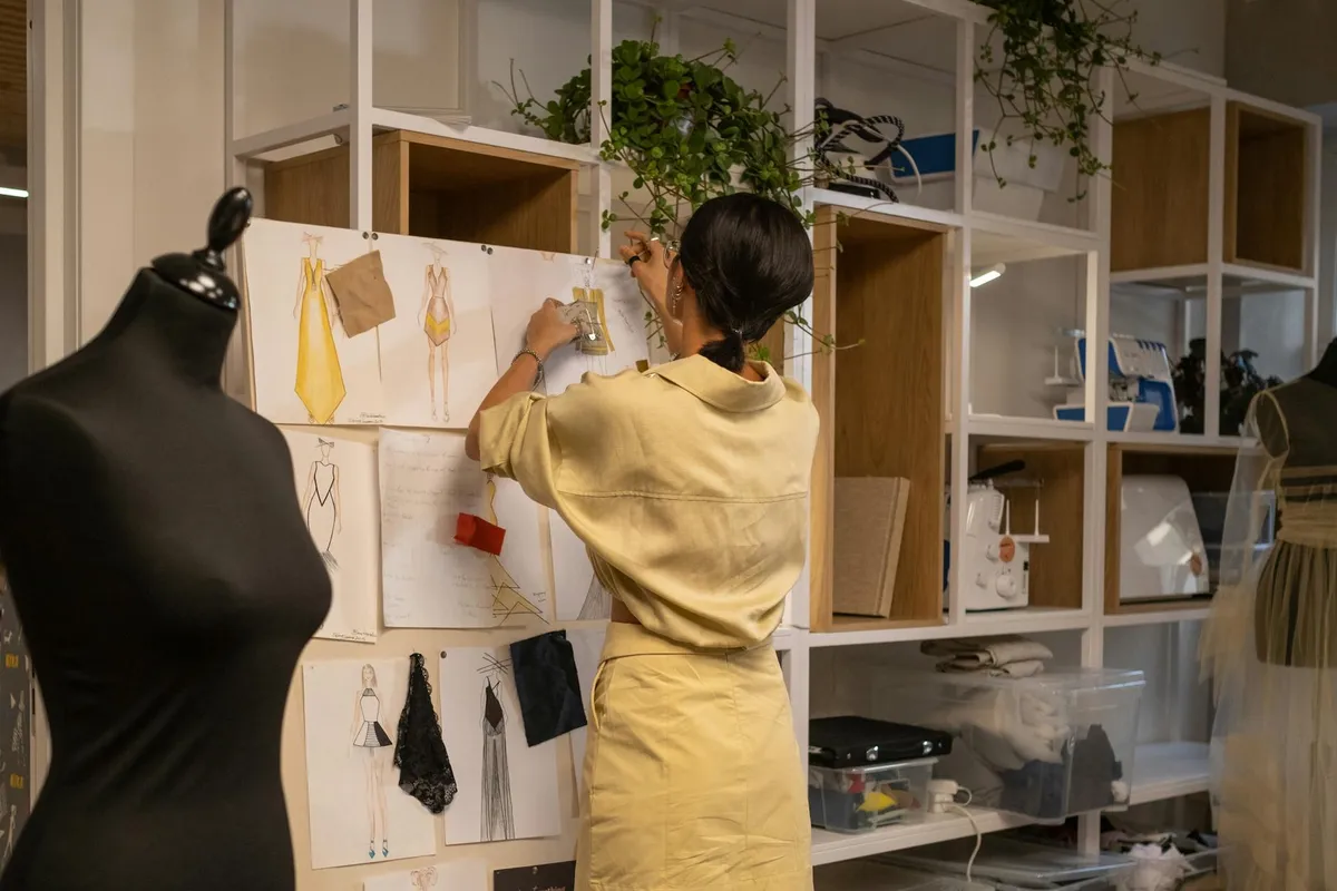 In a fashion design studio, a person in a light yellow outfit pins fabric swatches and sketches to a wall, with a mannequin nearby.