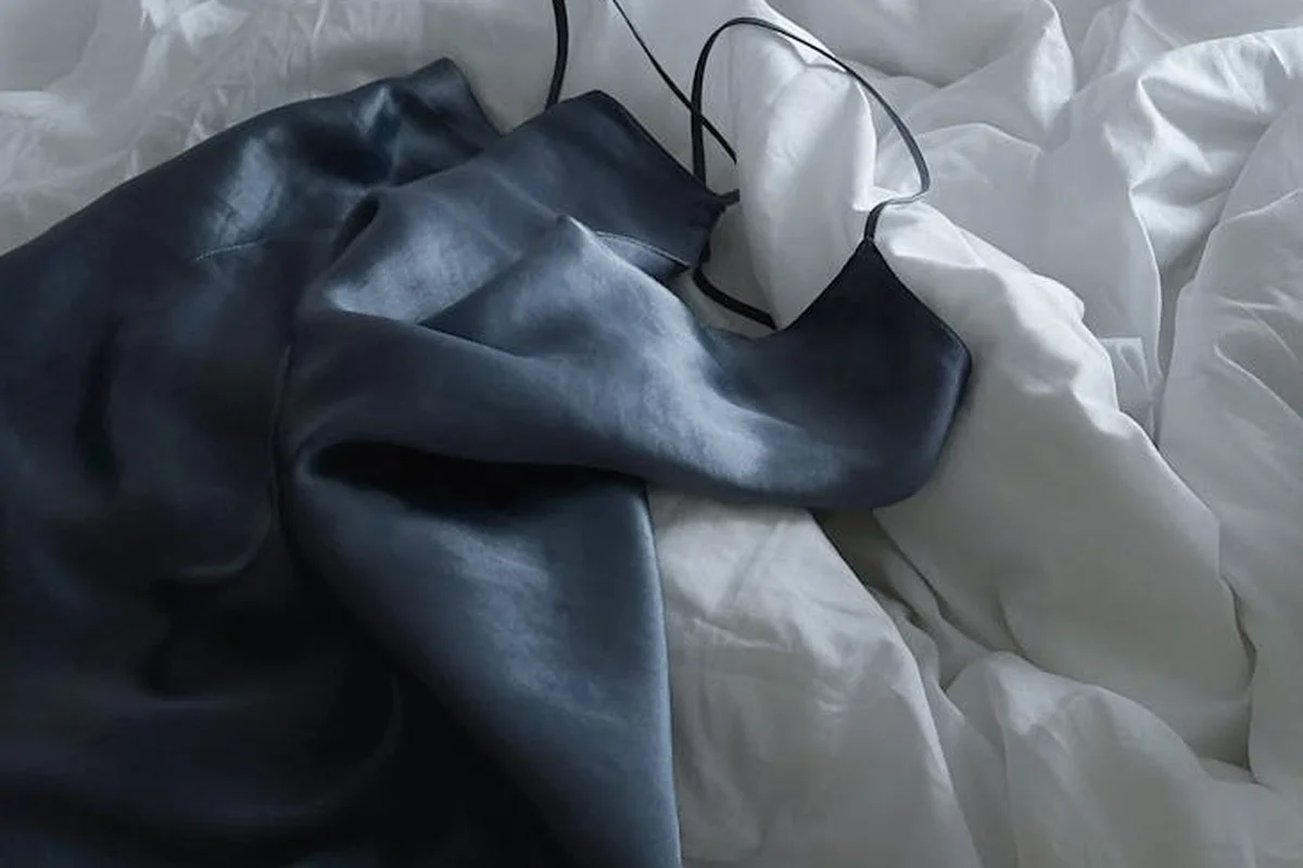 Close-up of dark blue satin-like fabric draped over white bed linens, highlighting smooth sheen and soft folds.