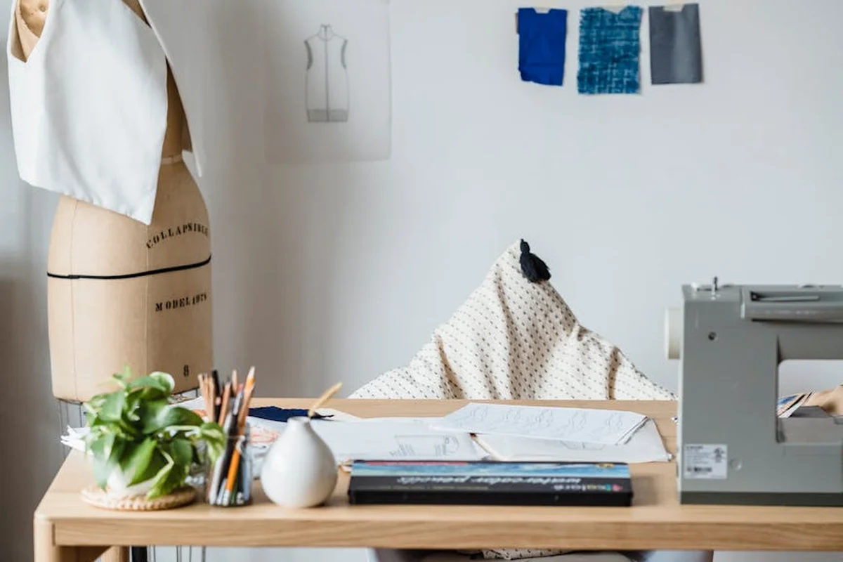 A bright sewing studio workspace featuring a dress form on the left, a wooden table with books and tools, and a sewing machine in the background, with fabric swatches on the wall.