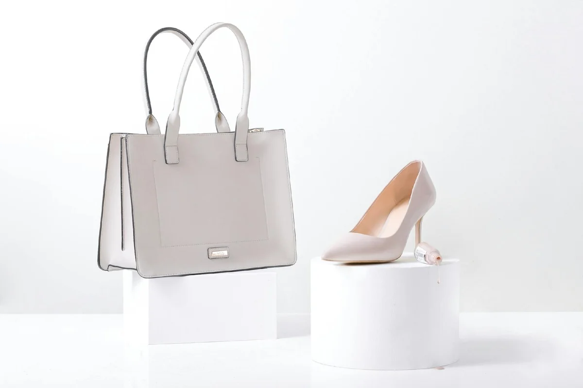Beige handbag and a matching beige high-heeled shoe displayed on white blocks against a white background