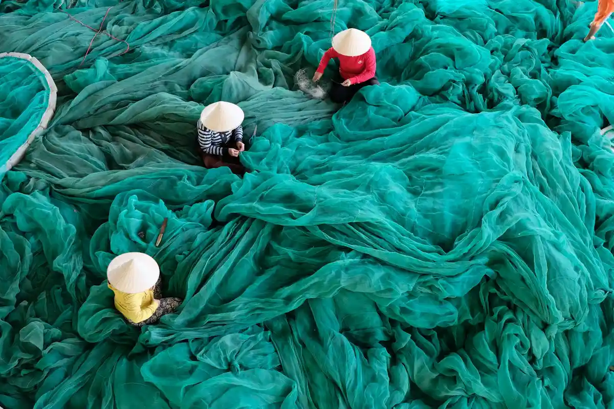 Several workers wearing white hats stand among large sheets of teal fabric spread across the workshop floor.