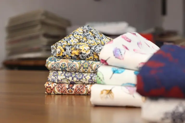 Stack of folded patterned fabrics on a wooden table, representing home textiles such as curtains and bedding.