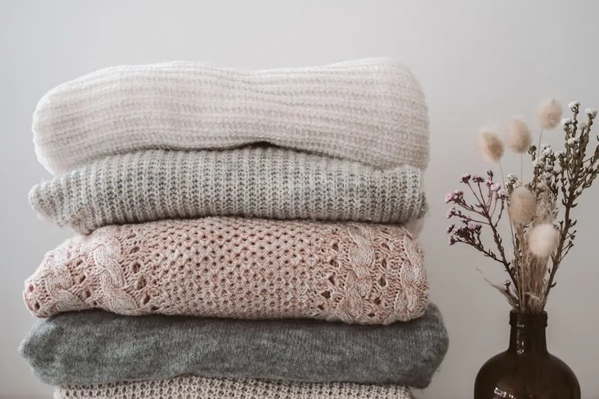 A stack of four folded sweaters in cream, beige, pink, and gray tones, placed beside a brown vase holding dried flowers.