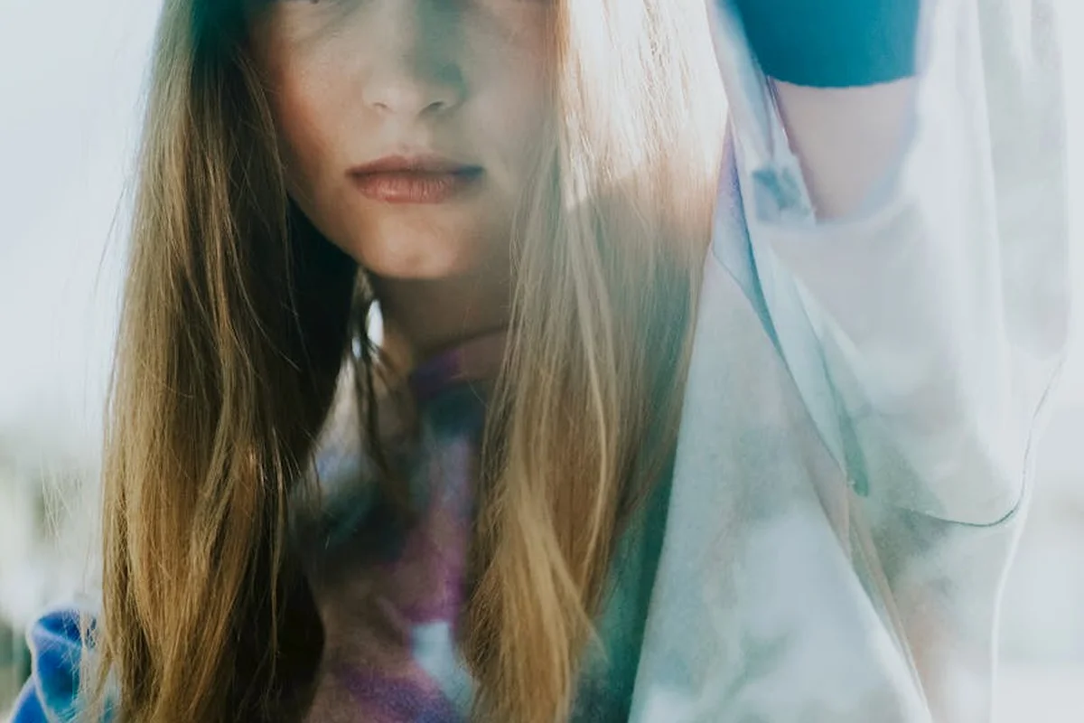 Close-up of a person with long hair wearing a white lab coat and blue gloves, with tie-dye fabric visible, implying dyeing with food coloring.