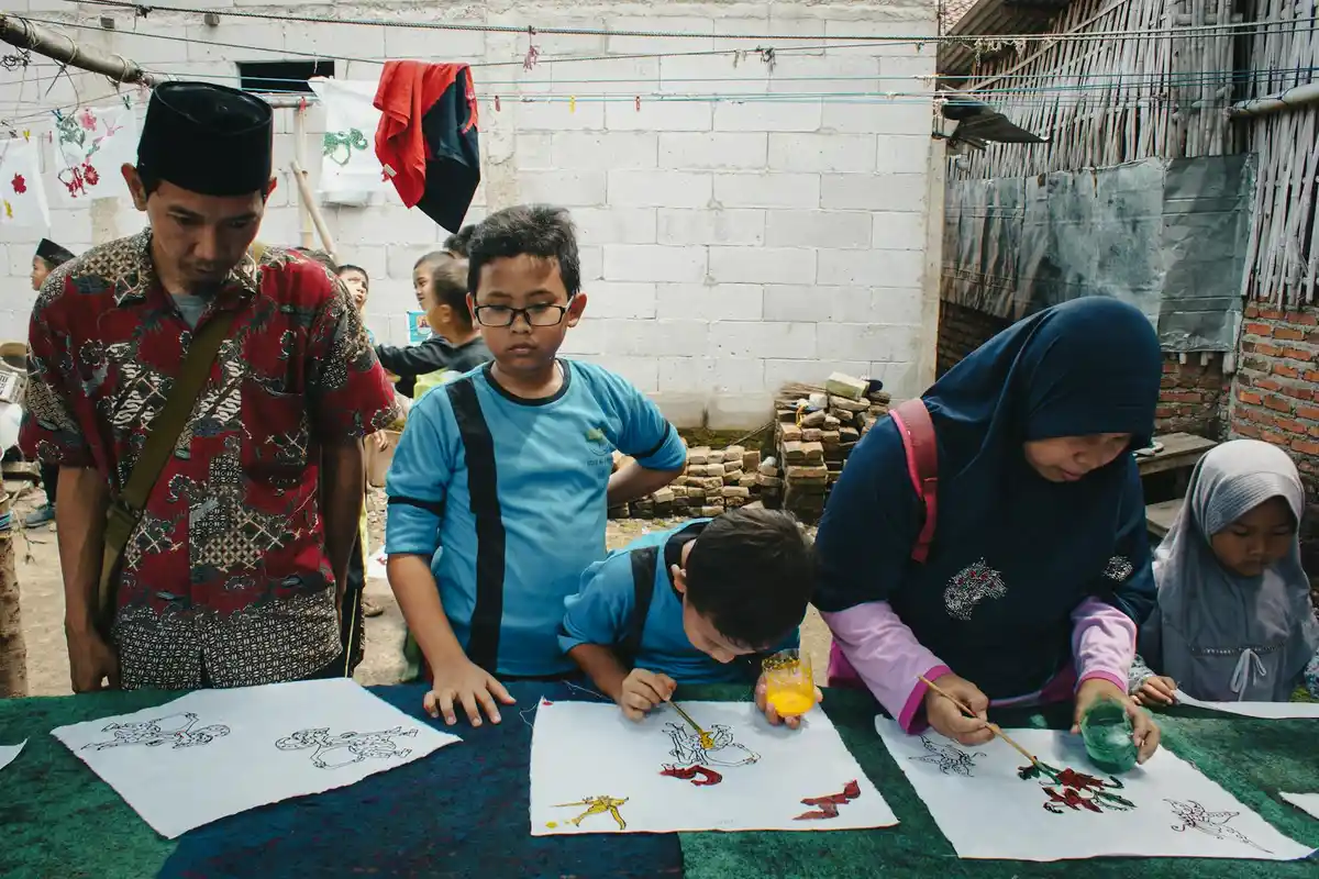Group of people, including children and adults, seated around a table crafting with paper and markers in a casual workshop setting.