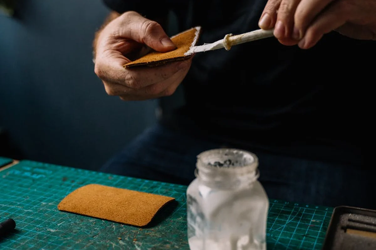 Hands apply glue from a syringe-like applicator onto a small fabric swatch on a green cutting mat, with a jar of glue nearby.