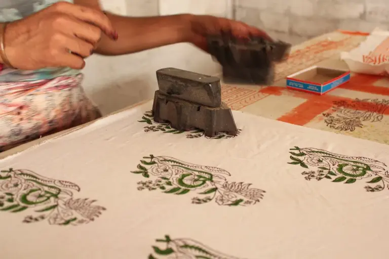Person wearing paint-splattered clothing applies dye to fabric with a stamping block on a patterned tabletop.
