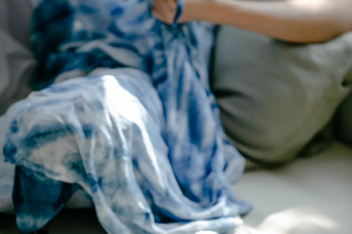 A person sits on a couch with blue and white hand-dyed fabric draped over their lap, illustrating a dyeing setup without a washing machine.
