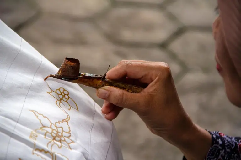 Close-up of a hand applying dye to white fabric using a wooden stamp-like tool, creating gold decorative patterns.