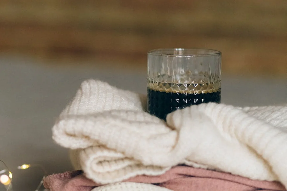 A stack of plush white knit fabrics with a pink layer beneath, and a glass in the background on a neutral surface.