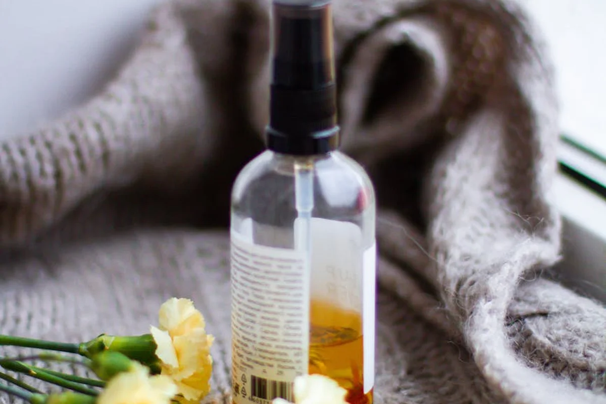 Clear spray bottle with a black nozzle sits on a soft beige knit fabric, with pale yellow flowers nearby, suggesting a DIY linen spray setup.