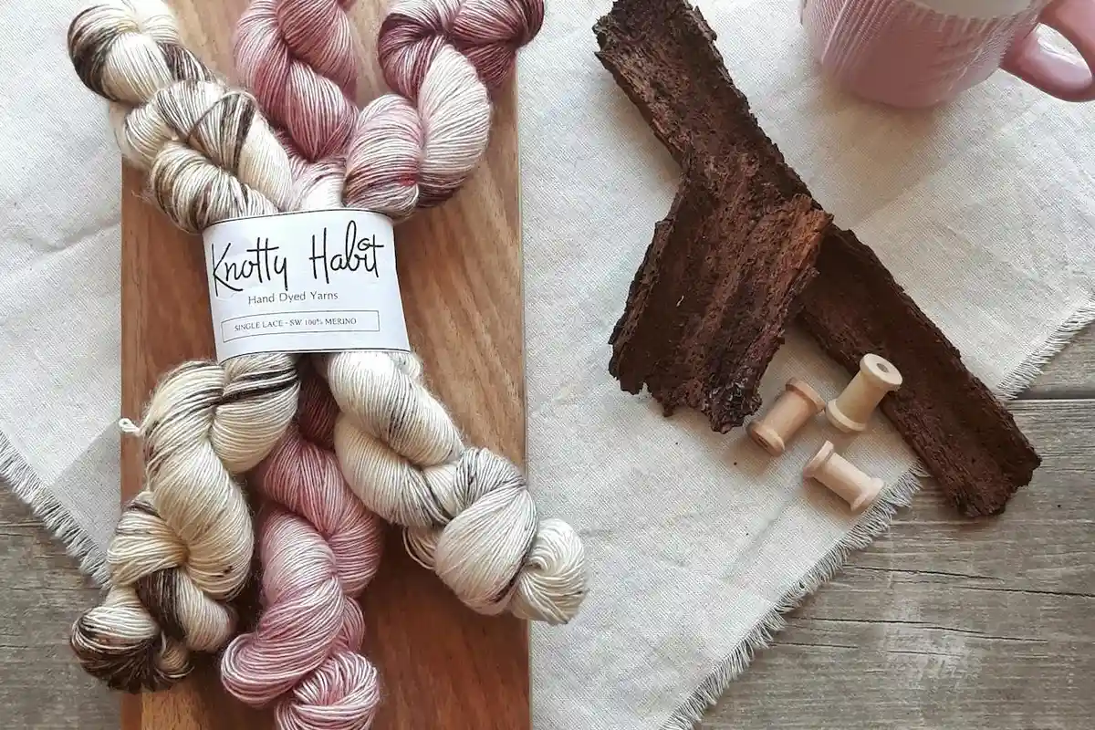 Two skeins of hand-dyed plant-fiber yarn on a wooden board, with a bark fragment and small spools nearby, showing natural pink and cream tones.