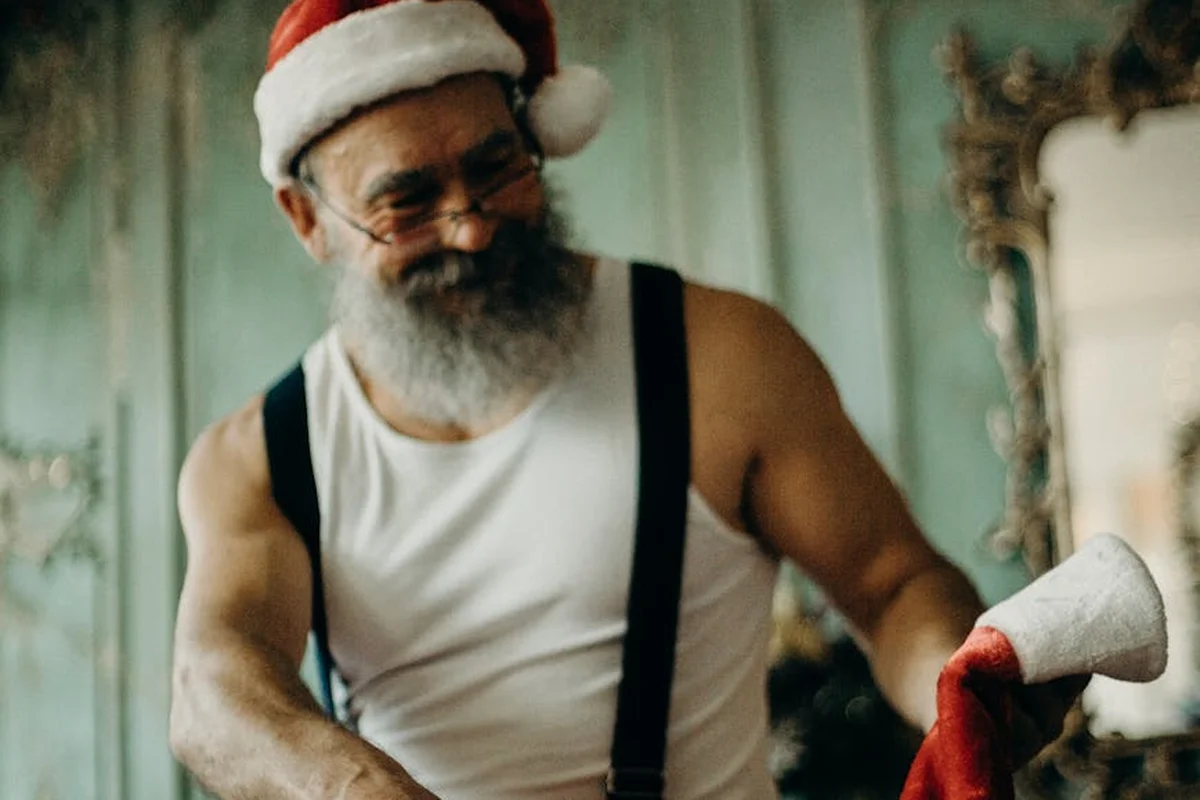 Smiling man in a Santa hat, white tank top and black suspenders, holding a red cloth in a vintage-looking room.