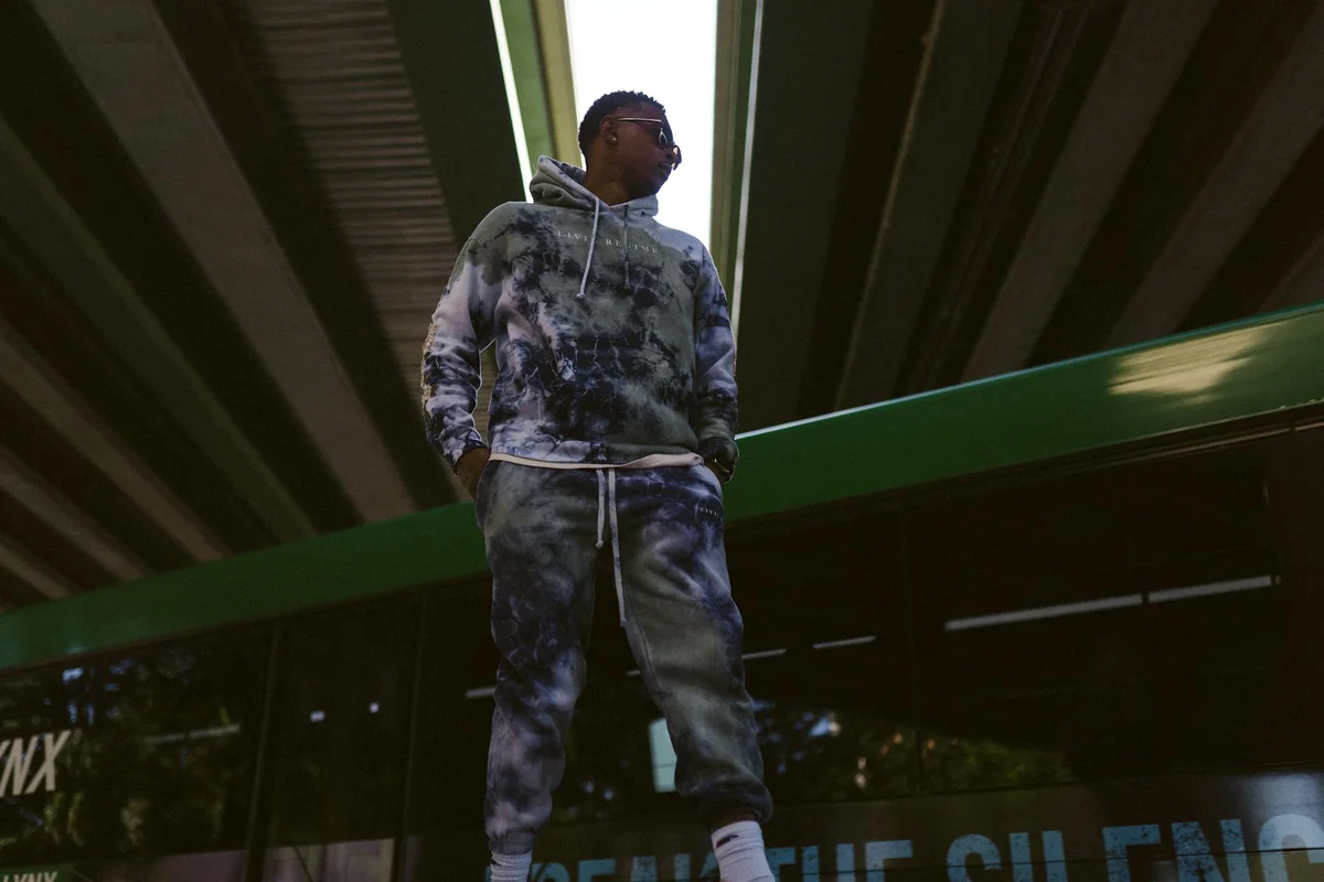 A person wearing a gray and black tie-dye hoodie and matching joggers stands under an overpass.