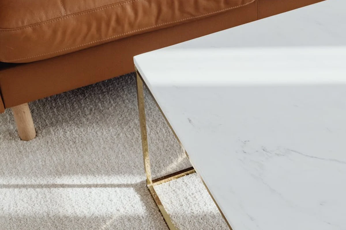 Close-up of a white marble table with gold legs beside a brown leather sofa on a light patterned carpet.