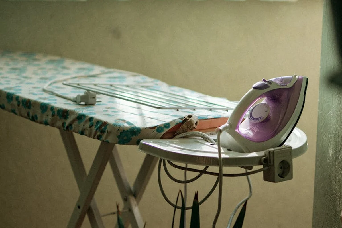 Ironing board with a purple electric iron resting on the iron rest, covered by a floral-patterned cover