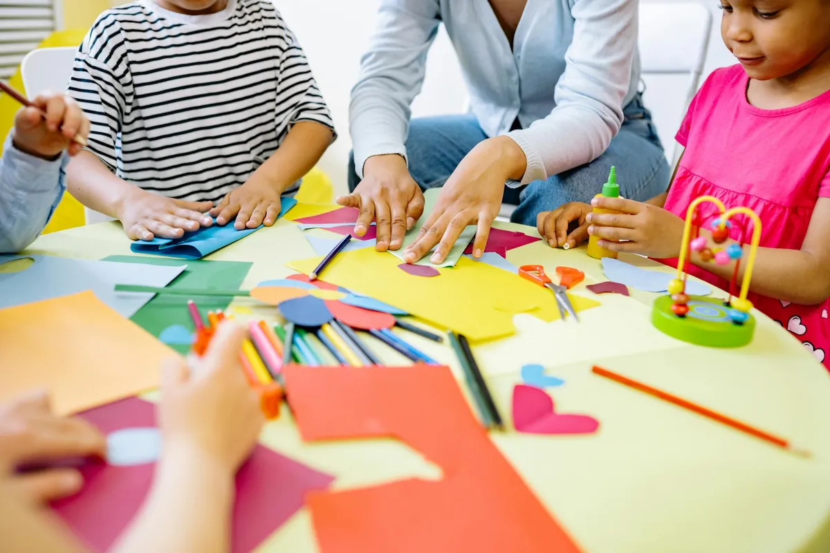 Children sit around a table making a craft project with colorful paper, glue, and scissors.