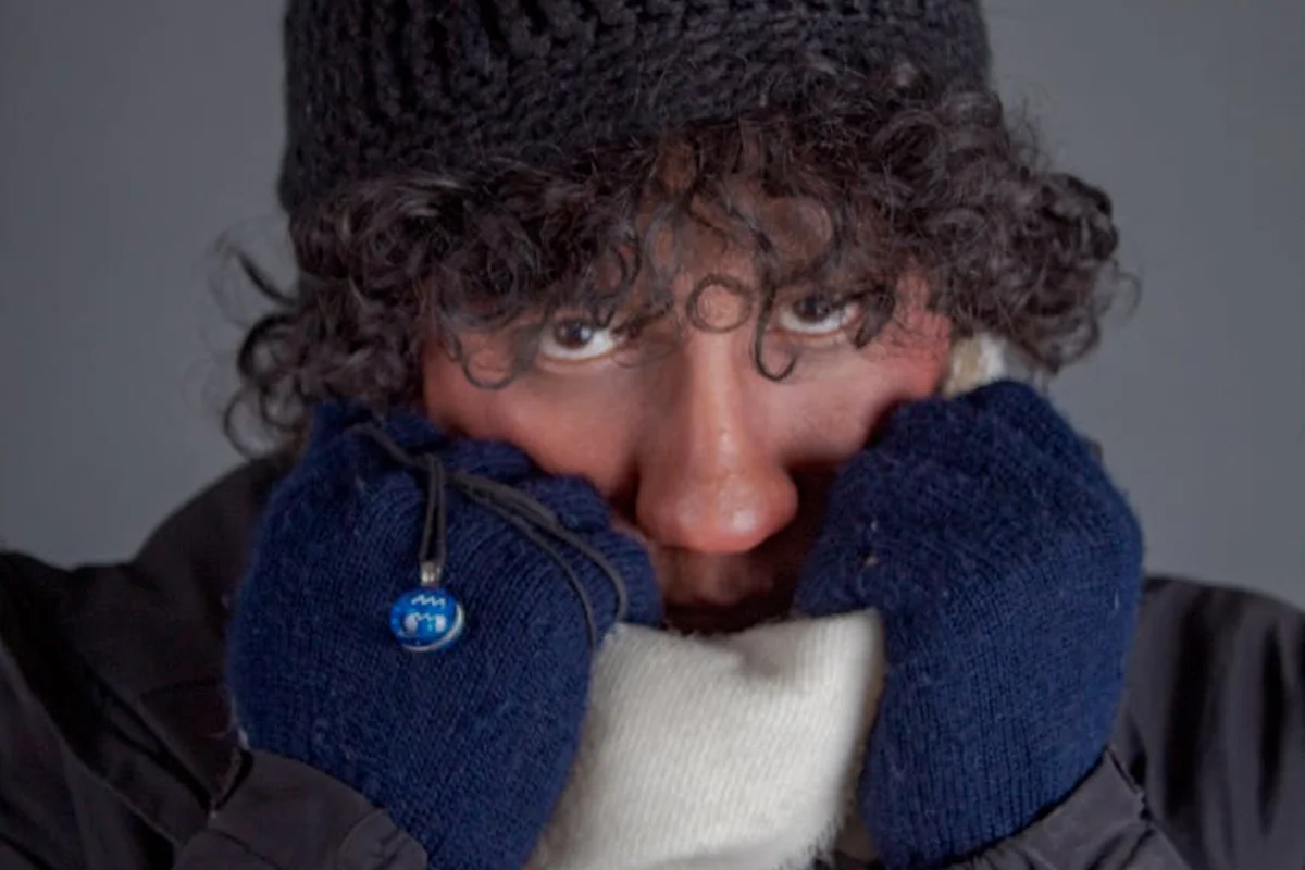 Close-up portrait of a person wearing a dark knit hat and blue wool gloves, with hands raised to cover their mouth and a wary expression.