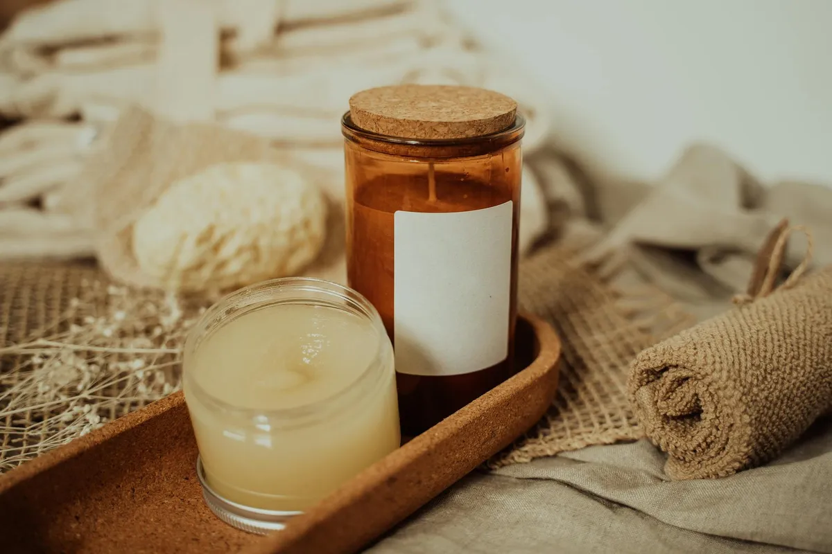 A small glass jar with pale cream, a brown corked bottle, and rolled burlap fabric on a wooden tray with linen textures in the background