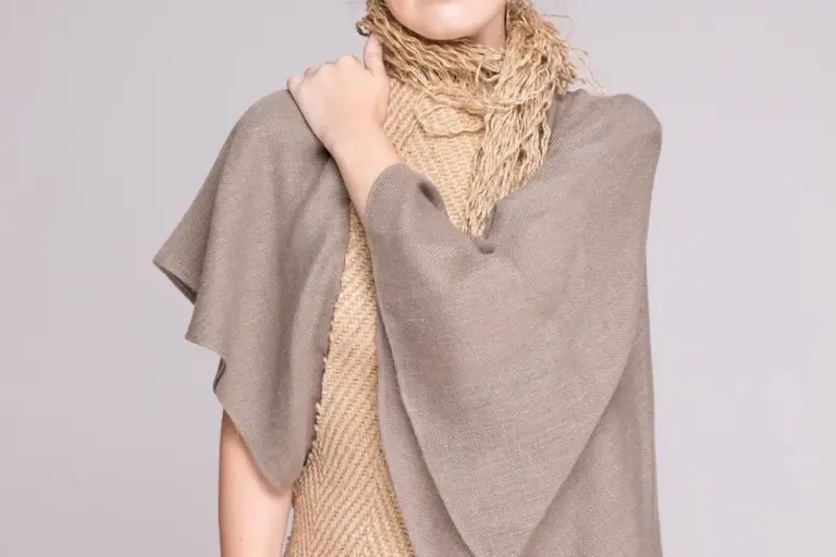 A person wearing a taupe linen shawl with a fringed scarf draped around the neck.